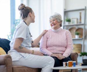This photo represents long-term care. It shows a senior woman in a pink sweater sitting next to a woman in white scrubs.