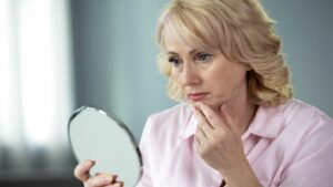 This photo depicts a senior woman looking at her skin in the mirror.