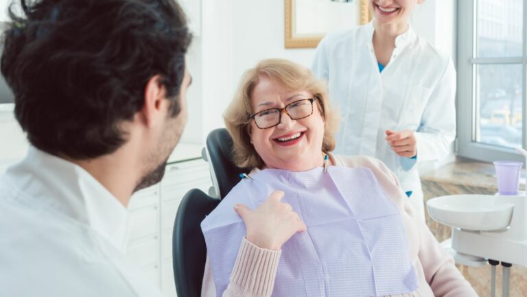 This image depicts a senior woman at the dentist. She is smiling at the doctor. The photo represents a senior receiving dentures.