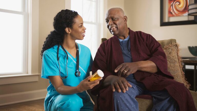 This image represents Medicare and long-term care insurance. A black woman sits with a black man. She is a nurse and is handing him medication.